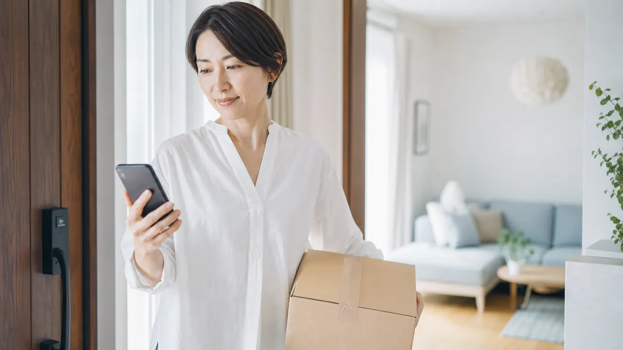 処方品の受け取りと再診予定を確認する様子 無地の荷物を受け取りスマートフォンで再診予定を確認する女性